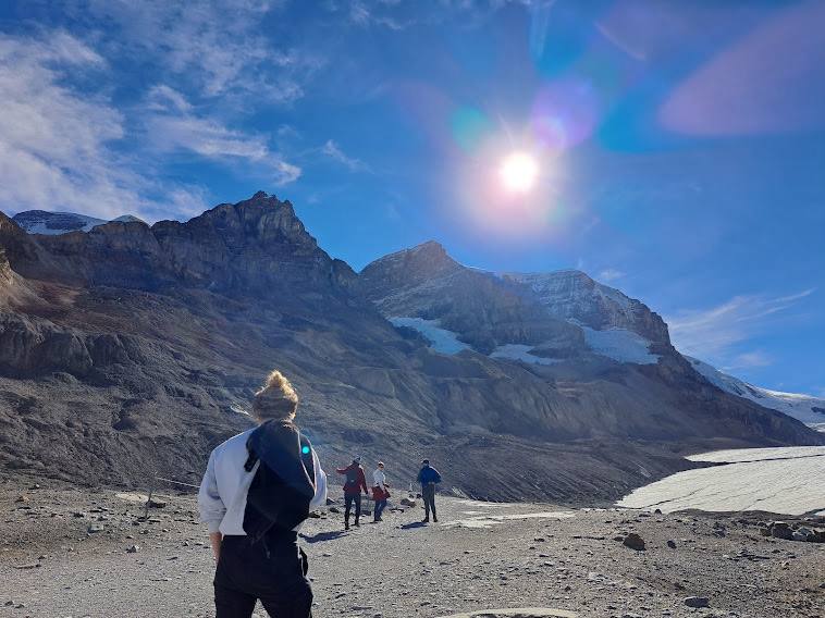 A group walking across glacial moraine towards the ice, below a jagged mountain ridge in bright sun